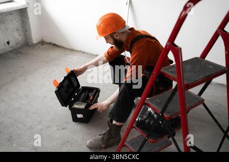 Male repairer. Handyman concept. Man in overalls opening toolbox sitting on ladder. Copy space Stock Photo