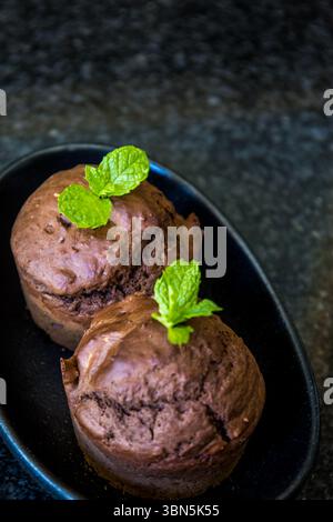 Two chocolate mint flavoured muffins, decorated with mint leaves, in a small, black elongated plate on a shiny stone table top. Stock Photo