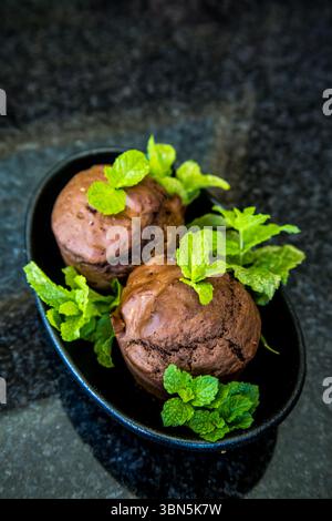 Two chocolate mint flavoured muffins, decorated with mint leaves, in a small, black elongated plate on a shiny stone table top. Stock Photo
