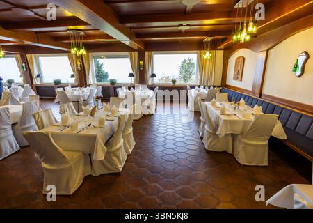 Empty restaurant dining room is ready for guests featuring white tablecloths, chair covers, and unique lighting fixtures in Kitzeck im Sausal, Styria, Stock Photo