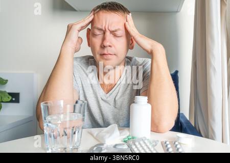 Sick man suffering from headache, migraine holding temples at home with medicine and water on table. Healthcare, cold and flu season, physical Stock Photo