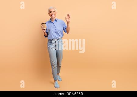 Stylish senior woman holding coffee cup, smiling warmly in casual wear against beige background Stock Photo