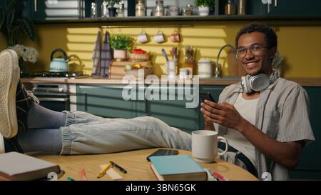 African American smiling man young guy student relax scrolling mobile phone gen z businessman freelancer putting legs on table studying at home Stock Photo