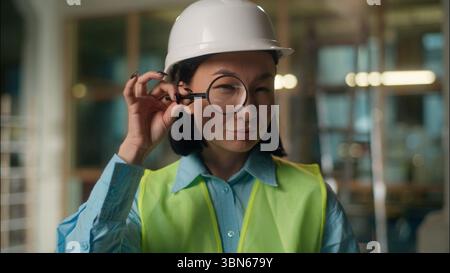 Smiling Asian builder Chinese girl architect engineer investigator Japanese Korean woman looking through magnifier vision test spy look with Stock Photo