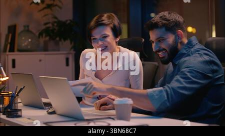 Multiracial businessman businesswoman Indian man Caucasian woman business partners co-workers looking at computer night evening office smiling Stock Photo