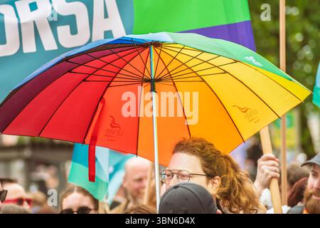 A smiling redhead woman walking out under rain with a rainbow umbrella amid the Pride Parade crowd. 28 June 2025 Stock Photo