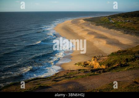 The ocean beach in Nazaré rests under calm weather, with gentle waves lapping the shore and soft light reflecting on the water. The scene feels peacef Stock Photo