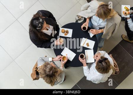Four female managers enjoy coffee and desserts during a lunch break, standing around a round table in a cafe or food court. Top-down aerial perspectiv Stock Photo
