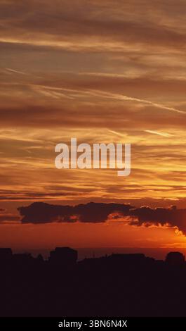 Golden sunset glows behind light clouds above a calm, silhouetted skyline, with soft transitions through orange, blue, and gray Stock Photo
