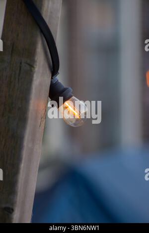 A glowing filament bulb hangs from a black cord attached to a wooden pole, softly lit against a blurred background of vertical lines and muted tones, Stock Photo