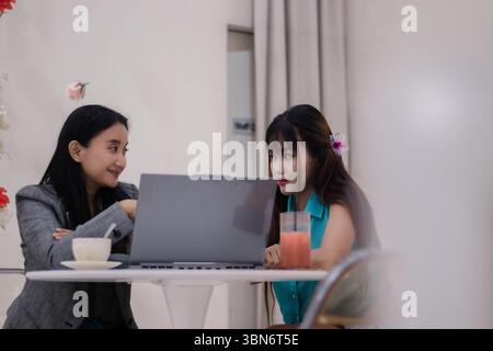 Two focused young women collaborating on a project in a cafe, reviewing data and sharing ideas on a laptop, drink included. Teamwork and business disc Stock Photo