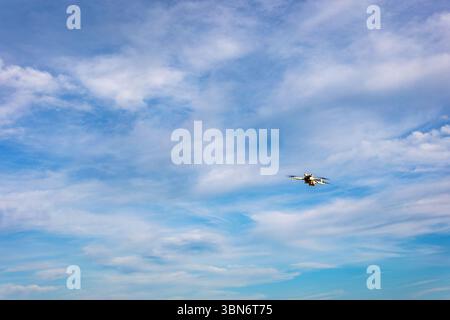 Flying drone in clear blue sky. A small drone captured mid-flight against a vivid blue sky with scattered clouds, symbolizing modern technology and Stock Photo