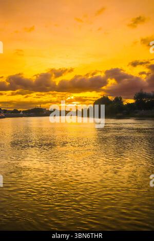 Vertical twilight view of golden sunset over river, shimmering reflections and tree silhouettes under cloudy sky, peaceful mood. Stock Photo