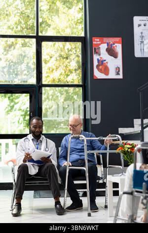 Black medic doing x ray scan interpretation with elderly man in the lobby, discussing about recovery steps and medication. Senior patient with reduced mobility uses a waling frame, evaluation. Stock Photo