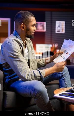 Young freelancer accountant doing math calculations using financial graphs on papers, working from his cozy home. Self employed person comparing business economic figures on files. Stock Photo