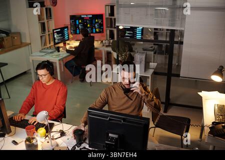 Young adult Caucasian man sitting at desk talking on phone while using computer, young adult Asian man typing on keyboard, woman working at computer in background in scam call center Stock Photo