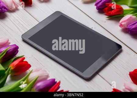 Grey tablet is lying flat on whitewashed table in flat design, surrounded by colorful tulip blooms Stock Photo