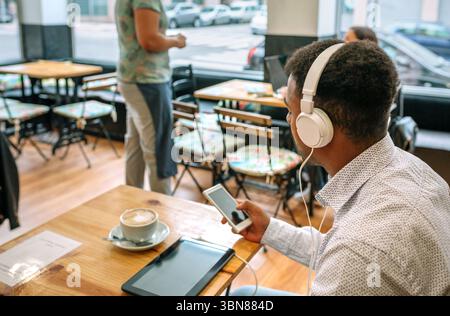 Unrecognizable young businessman listening music with headphones and using smartphone in cafe Stock Photo