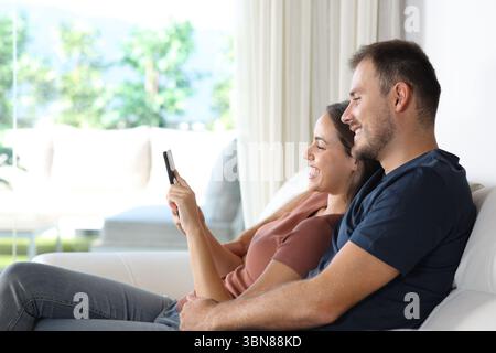 Profile of a happy couple checking mobile phone together in a house interior Stock Photo