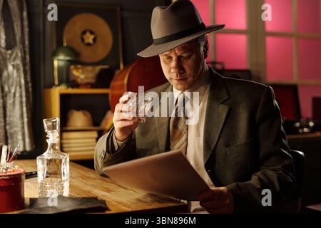 Middle aged Caucasian man wearing suit and fedora sitting at table reading sheet music while holding glass of whiskey, upright bass and jazz instruments visible in background Stock Photo