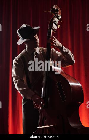 Caucasian young adult man wearing hat playing double bass on stage with red curtain background, focused expression, hands positioned on strings and neck of instrument Stock Photo