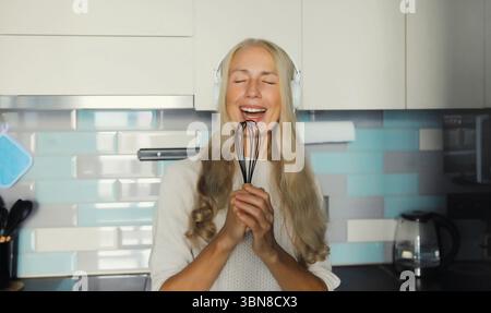 Happy cheerful mature woman singing with whisk listening to music with wireless headphones having fun in the kitchen at home in the morning Stock Photo