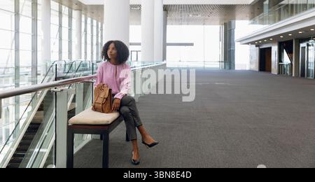 African American woman sitting on padded bench by escalator at expo with tan backpack, copy space Stock Photo