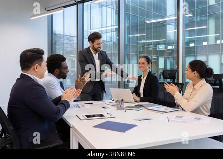 A diverse team celebrates a colleague's success in a modern office setting. Colleagues applaud and congratulate each other. Stock Photo