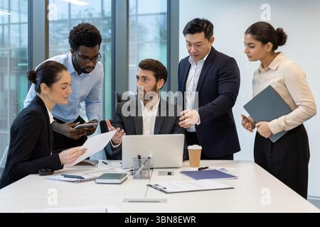 A diverse team of business professionals collaborating on a project in a modern office setting, using a laptop and documents. Stock Photo