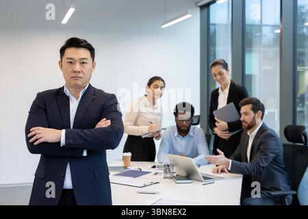 An Asian businessman stands with arms crossed in front of a diverse team in a modern office setting, focused on collaboration and teamwork. Stock Photo