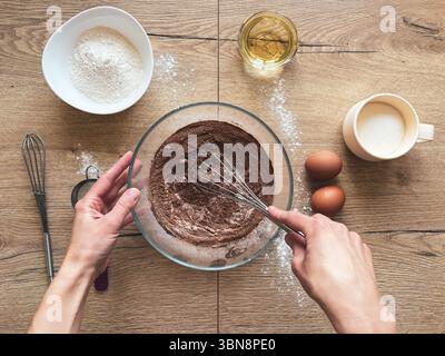 Female hands mixing dry ingredients for chocolate cake with a metal whisk. Homemade baking. Ingredients laid out on a wooden table Stock Photo