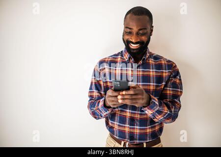 Happy black man in cozy shirt shirt standing in front of wall and messaging on smartphone. Stock Photo