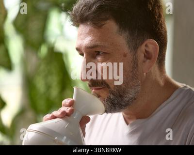 Man using steam inhaler for respiratory health indoors with green plant background. Aging with care Stock Photo