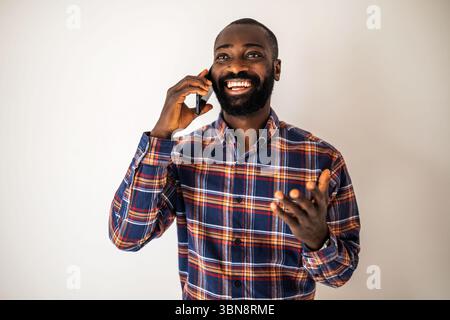 Cheerful black man in cozy shirt standing in front of wall and talking on mobile phone. Stock Photo