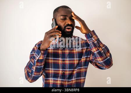 Anxious and tired black man in cozy shirt standing in front of wall and talking on mobile phone. Stock Photo