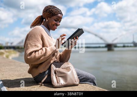 Young black woman is relaxing outdoor by the river. She is using digital tablet. Stock Photo