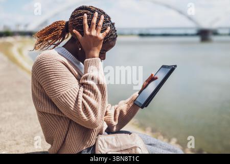 Young anxious black woman is using digital tablet. She is sitting by the river in the city. Stock Photo
