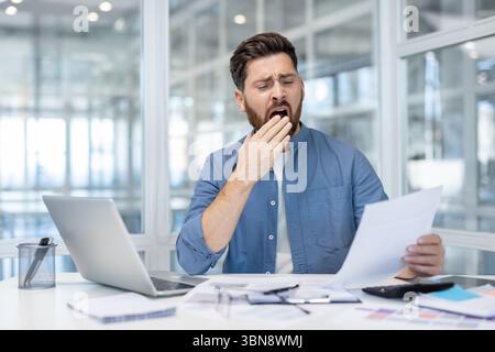 A bored businessman yawns while reviewing documents at his desk, showing signs of fatigue and exhaustion. Stock Photo