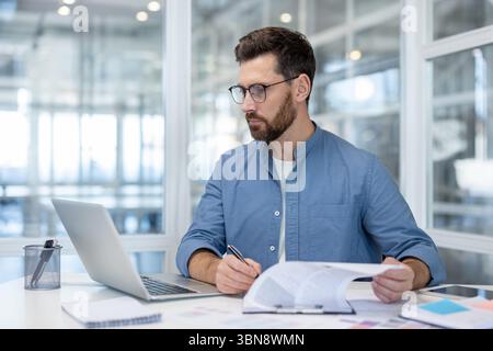 A professional man, seated at a desk, engrossed in paperwork, laptop, and working in office. Stock Photo