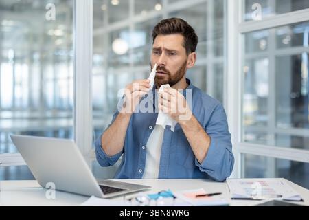 A man in an office setting administers nasal spray while using a tissue. A laptop and documents are on the desk. Stock Photo