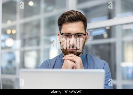 A thoughtful man with a beard wearing glasses looks intently at a laptop screen, hands clasped, in an office setting. Stock Photo