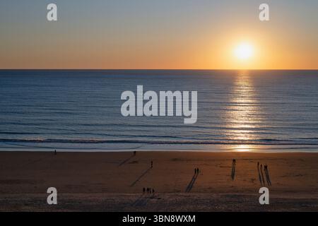 The sun sets on the horizon, painting the sky in shades of orange and gold over the wide, open beach in Cadiz, Spain Stock Photo