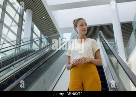Woman standing on descending escalator in office lobby wearing blouse, yellow pants under skylight Stock Photo