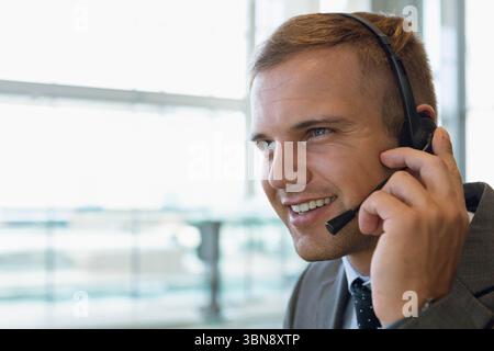 Man in suit and tie smiling, speaking into headset at call center near window, copy space Stock Photo