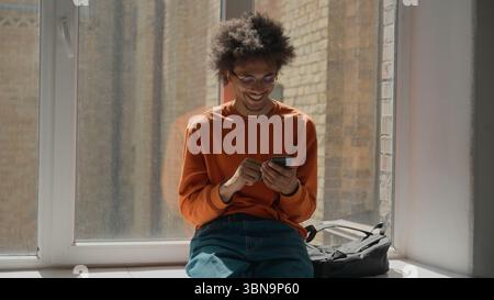 African American student guy using mobile phone relaxing near window in corridor university college school smartphone app chatting smiling happy Stock Photo