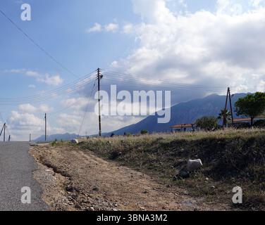 Cyprus, A rural landscape with a dirt road running through it. The road is flanked by power lines and poles, and a few houses are visible in the distance. The sky is a clear blue with fluffy white clouds. In the background, a range stretches across the horizon, with a mix of green and brown vegetation. The foreground features a grassy area with some rocks and a small, light-colored rock. An artist's eye & imagination shaped Stock Photo