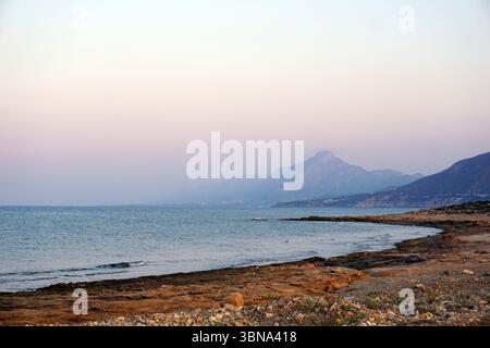 Cyprus, Kyrenia Mountains, Pentadaktylos, A serene coastal landscape. The foreground features a rocky shore with a mix of brown and tan colors, and a few small rocks scattered on the ground. The calm, light blue-green sea stretches out to the horizon, where it meets a hazy, light blue-gray sky. In the distance, a mountain range with a prominent peak rises against the horizon. The mountains are a mix of brown and gray tones. The overall color palette of the image is muted, with soft pastel hues dominating the sky and sea. An artist's eye & imagination shaped Stock Photo