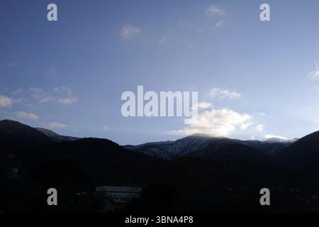 traveling in Cyprus, Troodos Observatory, A serene mountain range under a clear blue sky. The mountains, covered in snow, stretch across the horizon, their peaks reaching towards the sky. The sky is a light blue with a few scattered white clouds. In the foreground, a dark silhouette of a settlement is visible, with a few buildings and trees. The image is taken from a high vantage point, providing a panoramic view of the mountains and the settlement below. An artist's eye & imagination shaped Stock Photo