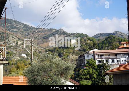 Cyprus village, A serene mountainous landscape with a clear blue sky dotted with fluffy white clouds. The mountains are covered in a mix of green and brown vegetation, with some areas appearing dry or barren. In the foreground, a cluster of buildings with red tile roofs and white walls are nestled on a hillside, with a mix of trees and shrubs in the foreground. Utility poles and power lines are visible in the mid-ground, and a small white house is perched on a hilltop in the background. The perspective of the image is from a low angle, looking up at the mountains, creating a sense of grandeur Stock Photo