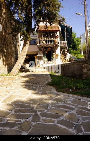 Cyprus, Edremit, A quaint street scene with a stone path made of irregularly shaped stones, leading towards a white building with a red tile roof. The building has a balcony adorned with various pots and vases, and a small shop or store is visible on the ground floor. A tree stands on the left side of the path, casting a shadow on the ground. The sky is a clear blue, and power lines run along the street. A stone wall is visible on the right side of the image, and a streetlight is present on the right side. The building has a small, dark-colored awning or covering over the entrance. An Stock Photo